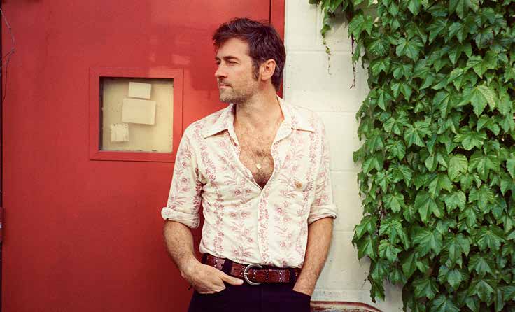 John Craigie standing beside a red door and a wall covered in green ivy.
